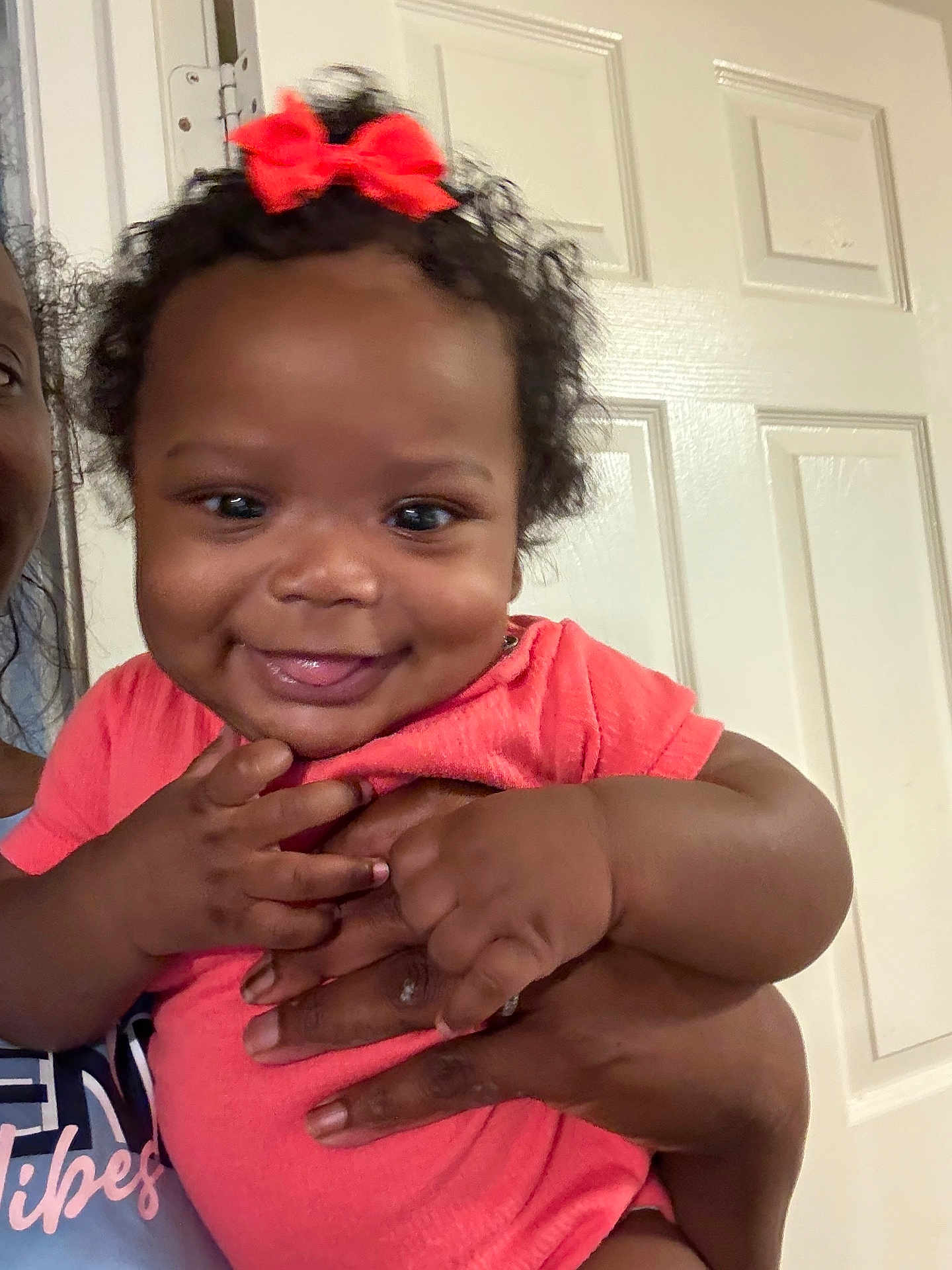 Curcedes is registered to the contest to win money with this photo: baby, child, smiling, red_bow, curly_hair, hand, adult_hand, indoor, door, portrait, closeup, happy, person, cute, infant, skin, clothing, pink_shirt, holding, face