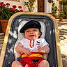 baby, child, smiling, stroller, beret, clothing, outdoor, potted_plants, flowers, sign, door, courtyard, sunlight, shadow, happy, person, red_clothing, white_shirt, yellow_belt, spain