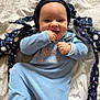 baby, infant, smile, onesie, blue_clothing, knit_hat, hand_in_mouth, blanket, bedding, jacket, pattern, polka_circles, portrait, cute, lying_down, happy, fingers, face, indoors, bedroom