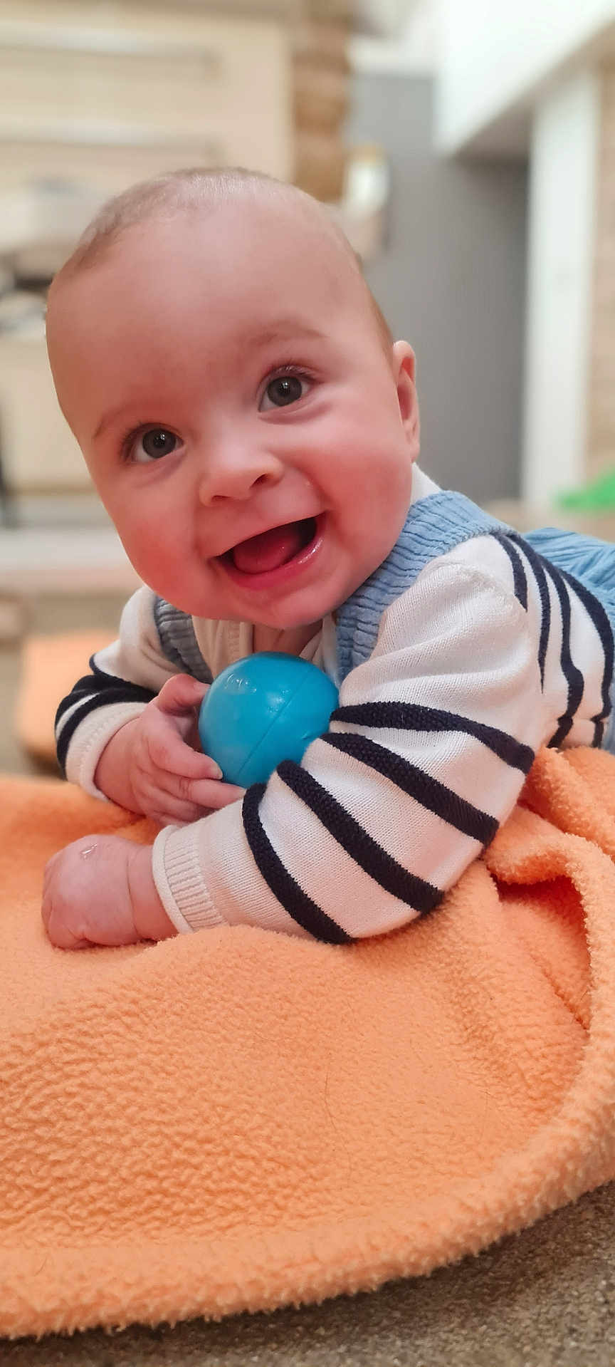 Raphaël participe au concours pour gagner de l'argent avec cette photo : baby, infant, smiling, blue_ball, orange_blanket, striped_sweater, lying_on_tummy, hands, close_up, portrait, indoors, big_eyes, open_mouth, cheeks, soft_texture, cute, playful, toy, happy, nursery
