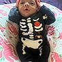 Amelia is registered to the contest to win money with this photo: baby, infant, child, skeleton_outfit, halloween, costume, orange_hat, cushion, floral_pattern, blanket, white_socks, cute, portrait, indoors, soft_lighting, hands_near_head, small_face, lying_down, black_clothing, holiday_theme