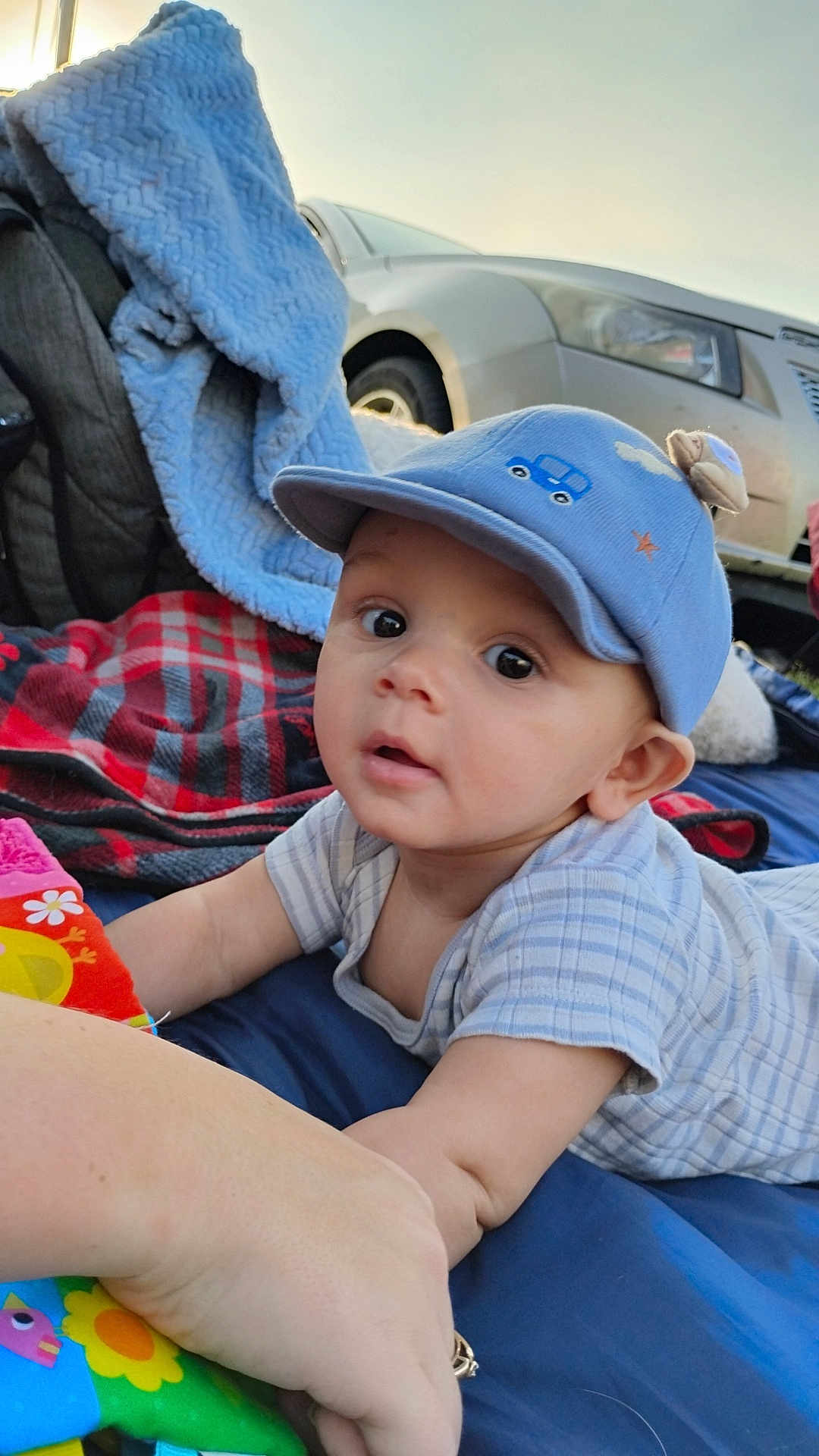 Milo is registered to the contest to win money with this photo: baby, child, blue_cap, blanket, colorful, curious, outdoor, car, face, wide_eyes, reaching, lying_down, plaid_blanket, soft_blanket, person, infant, close_up, casual, daytime, cute