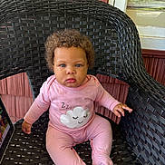 Tori joined the competition — help win amazing prizes! baby, child, curly_hair, pink_clothing, onesie, sitting, wicker_chair, porch, window, wooden_siding, face, serious_expression, indoor, person, cute, infant, cozy, closeup, hands, feet