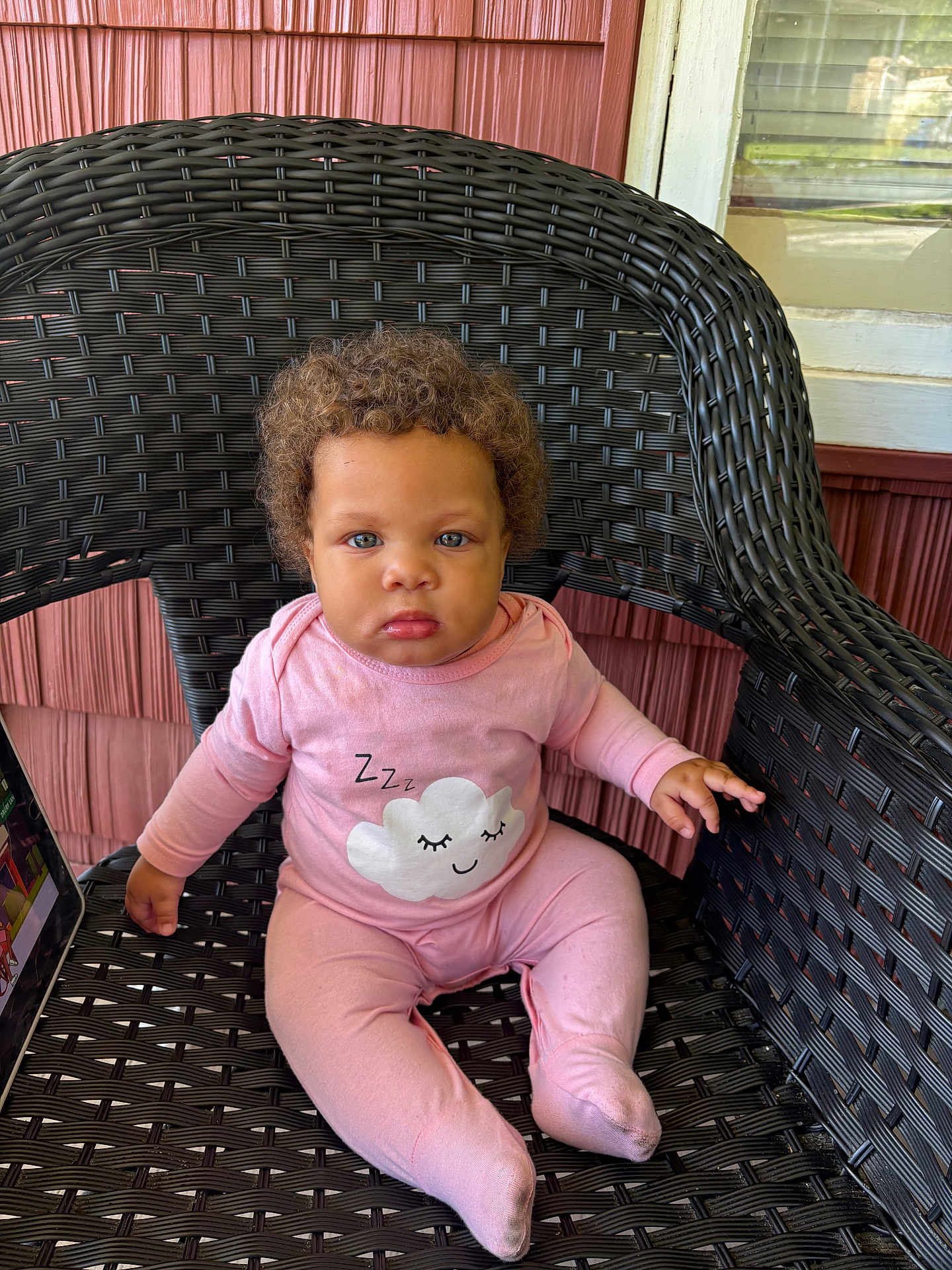 Tori joined the competition — help win amazing prizes! baby, child, curly_hair, pink_clothing, onesie, sitting, wicker_chair, porch, window, wooden_siding, face, serious_expression, indoor, person, cute, infant, cozy, closeup, hands, feet
