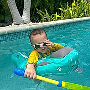 Conner is registered to the contest to win money with this photo: toddler, child, pool, float, sunglasses, water_toy, summer, outdoor, swimming, blue_water, greenery, palm_trees, sunny, baby, yellow_clothing, fun, vacation, leisure, playful, swimwear