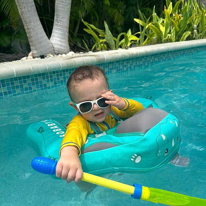 Conner is registered to the contest to win money with this photo: toddler, child, pool, float, sunglasses, water_toy, summer, outdoor, swimming, blue_water, greenery, palm_trees, sunny, baby, yellow_clothing, fun, vacation, leisure, playful, swimwear