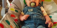 Matteo is registered to the contest to win money with this photo: baby, pacifier, denim_overalls, baby_rocker, indoor, child, infant, clothing, foot, face, person, comfort, colorful, pattern, furniture, cute, casual, relaxed, young_child, seated