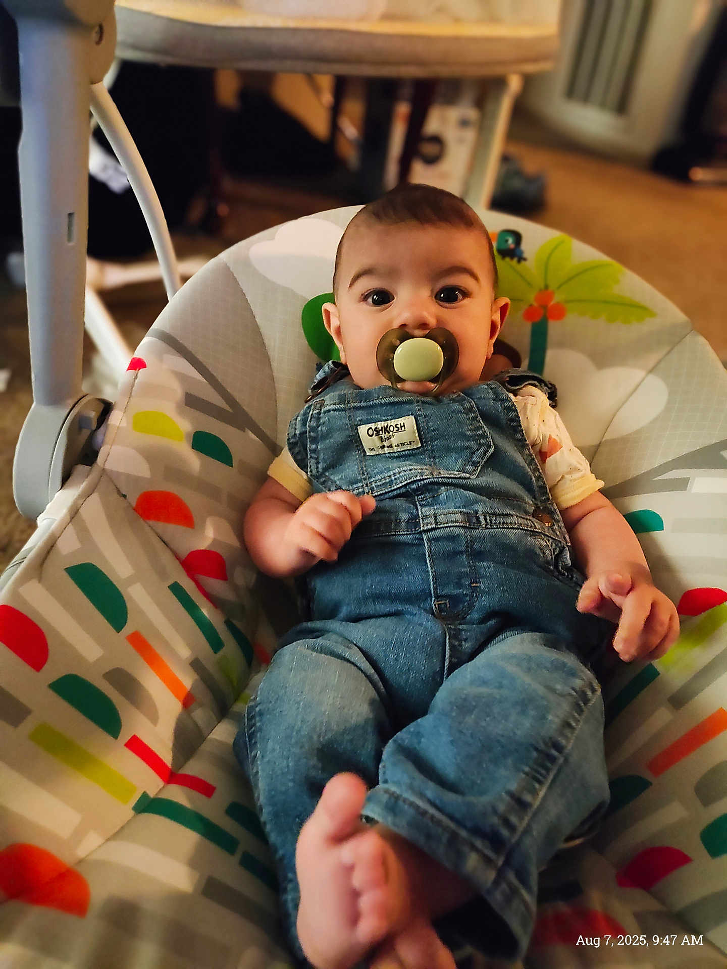 Matteo is registered to the contest to win money with this photo: baby, pacifier, denim_overalls, baby_rocker, indoor, child, infant, clothing, foot, face, person, comfort, colorful, pattern, furniture, cute, casual, relaxed, young_child, seated