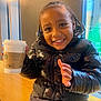 Carson joined the competition — help win amazing prizes! child, smiling, indoor, puffer_jacket, braided_hair, starbucks_cup, coffee, table, lamp, window, happy, portrait, casual_clothing, seated, young, person, warm_lighting, wooden_surface, black_jacket, cute