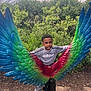 child, boy, wings, sculpture, outdoor, greenery, trees, adidas, clothing, playful, posing, park, daylight, casual, smile, colorful, art, nature, young, fun