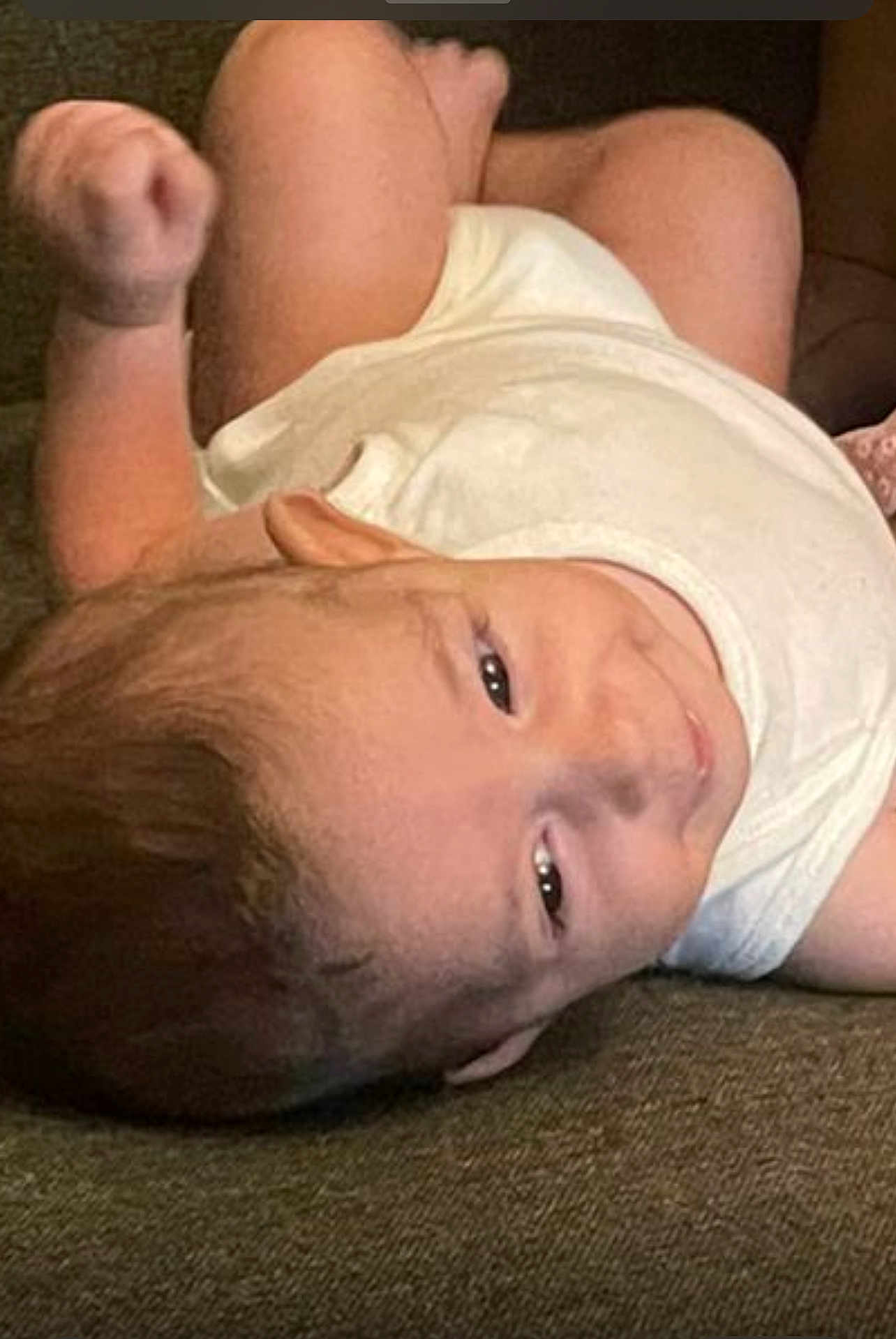 Yahya is registered to the contest to win money with this photo: baby, infant, child, white_clothing, sleeveless_shirt, lying_down, curious_expression, raised_arm, skin, head, face, eyes, smile, hair, person, indoors, couch, fabric, texture, relaxed