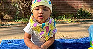 Eithan is registered to the contest to win money with this photo: baby, bib, blue_blanket, brick_wall, casual_clothing, child, colorful, curious, cute, daytime, hat, infant, nature, outdoor, person, plastic_eggs, playful, sitting, sunlight, toys