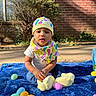 baby, bib, blue_blanket, brick_wall, casual_clothing, child, colorful, curious, cute, daytime, hat, infant, nature, outdoor, person, plastic_eggs, playful, sitting, sunlight, toys