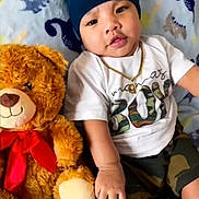 Ignatius is registered to the contest to win money with this photo: baby, teddy_bear, child, hat, white_tshirt, gold_chain, camouflage_shorts, blanket, dinosaur_pattern, stuffed_animal, red_bow, cute, indoors, smiling, sitting, soft_toy, infant, portrait, cozy, happy