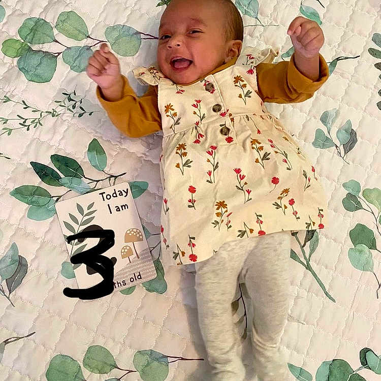 Sophia is registered to the contest to win money with this photo: baby, babylaughing, bodypart, clothing, coat, dress, face, finger, furniture, hand, happy, head, pants, person, photography, portrait, quilt, sitting, smile, text