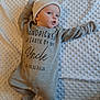Albie is registered to the contest to win money with this photo: baby, infant, onesie, hat, blanket, lying_down, expression, face, child, cute, cozy, text, message, gray_clothing, soft_texture, indoors, portrait, newborn, sleepwear, warm