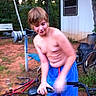 child, boy, shirtless, blue_shorts, bicycle, outdoor, exercise_equipment, building, window, grass, dirt, tree, motion_blur, summer, casual, young, active, play, person, nature