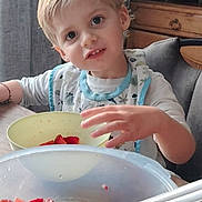 Lonaël a rejoint le concours — aidez-le/la à gagner de superbes lots ! bib, blonde_hair, bowl, child, clothing, curious, facial_expression, food, fruit, hand, indoor, kitchen, meal, paper_towel, person, sitting, spoon, strawberries, table, wooden_furniture