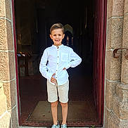 Gabin a rejoint le concours — aidez-le/la à gagner de superbes lots ! child, boy, smiling, white_shirt, shorts, sneakers, standing, doorway, stone_wall, entrance, casual_clothing, outdoor, daylight, young, portrait, happy, person, architecture, footwear, pose