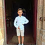 child, boy, smiling, white_shirt, shorts, sneakers, standing, doorway, stone_wall, entrance, casual_clothing, outdoor, daylight, young, portrait, happy, person, architecture, footwear, pose