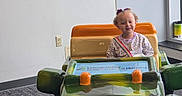 Monroe is registered to the contest to win money with this photo: child, girl, smiling, portrait, toy_car, toy_jeep, driving, hospital, indoor, playroom, vehicle, green_car, orange_car, rainbow_shirt, curly_hair, hair_bows, seated, happy, wall, carpet