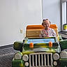 child, girl, smiling, portrait, toy_car, toy_jeep, driving, hospital, indoor, playroom, vehicle, green_car, orange_car, rainbow_shirt, curly_hair, hair_bows, seated, happy, wall, carpet