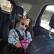 Monroe is registered to the contest to win money with this photo: child, toddler, car_seat, sunglasses, pacifier, sippy_cup, winter_coat, car_interior, seatbelt, bow, curly_hair, cup_holder, backseat, window, snow, road, travel, safety, relaxed, portrait