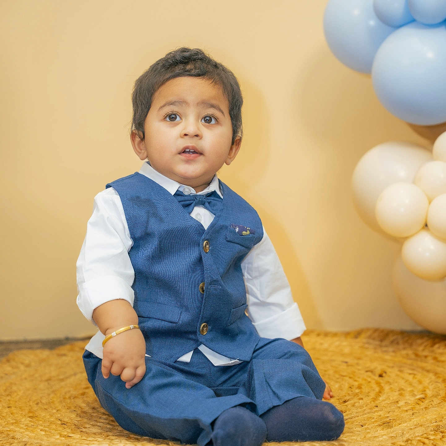 Venkata Avyukt joined the competition — help win amazing prizes! toddler, child, boy, blue_vest, bow_tie, white_shirt, sitting, indoor, balloons, decorations, curly_hair, curious_expression, woven_mat, portrait, cute, fashion, celebration, happy, floor, photography