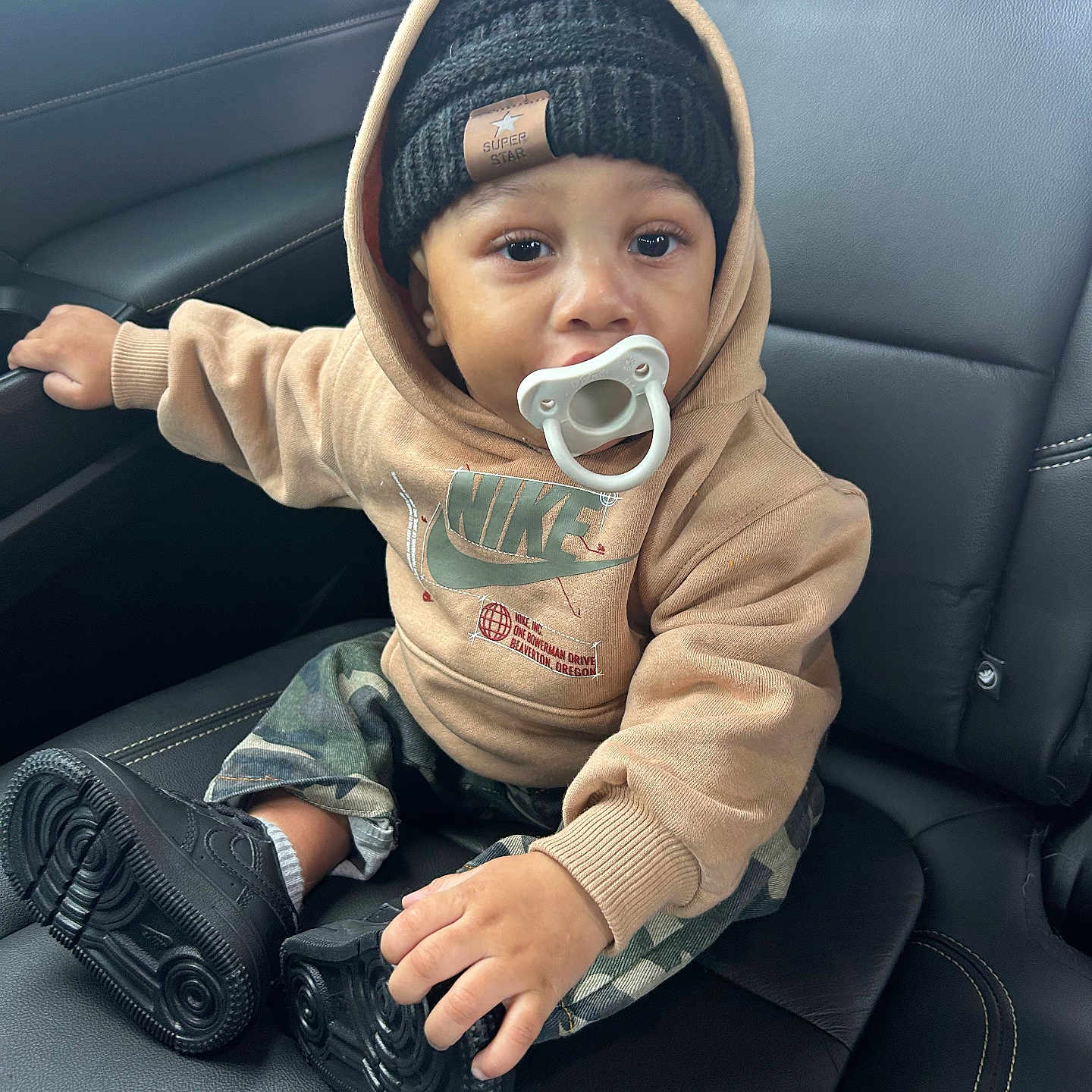 Shyne is registered to the contest to win money with this photo: baby, beanie, black_shoes, camo_pants, car_seat, casual, child, clothing, cute, face, footwear, headwear, hoodie, indoor, leather_seat, pacifier, portrait, sitting, toddler, young_child