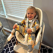 Oakley is registered to the contest to win money with this photo: toddler, child, high_chair, pajamas, sunflower_pattern, smile, indoor, window_blinds, happy, socks, furniture, floor_mat, person, seat_belt, light, wooden_leg, wall, carpet, home, cute