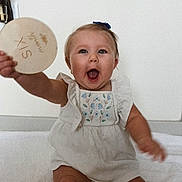 Maci joined the competition — help win amazing prizes! baby, child, infant, smiling, happy, sitting, blue_bow, plaque, wooden, celebration, milestone, white_dress, embroidered, cute, portrait, indoors, blanket, person, face, eyes
