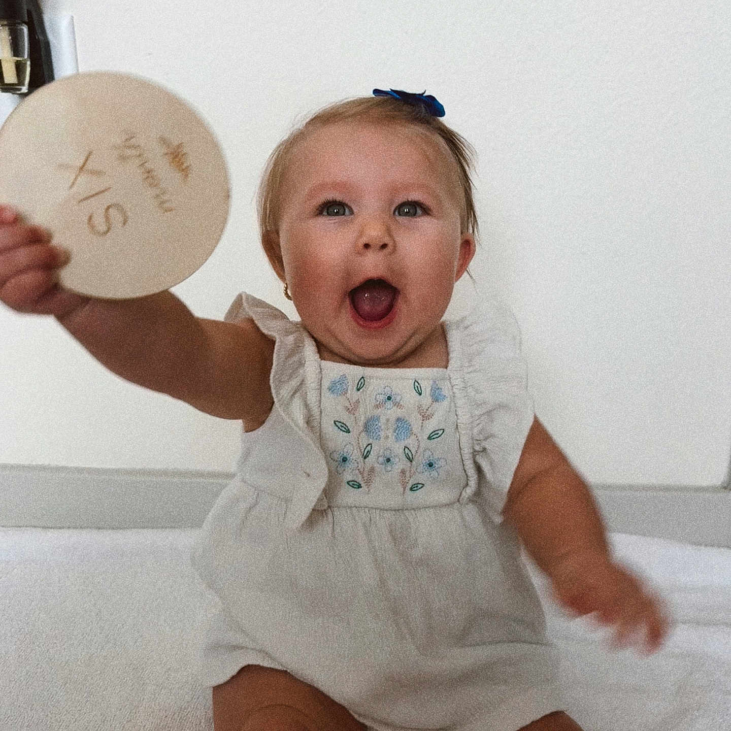 Maci joined the competition — help win amazing prizes! baby, child, infant, smiling, happy, sitting, blue_bow, plaque, wooden, celebration, milestone, white_dress, embroidered, cute, portrait, indoors, blanket, person, face, eyes