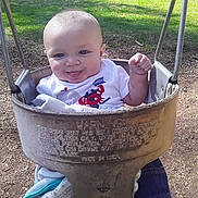 Taylon joined the competition — help win amazing prizes! baby, child, infant, playground, swing, smiling, tongue, fist, outdoor, grass, park, seated, portrait, white_shirt, red_shirt_graphic, safety_seat, playground_equipment, sunlight, shade, happy