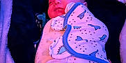 Kody is registered to the contest to win money with this photo: baby, sleeping, blanket, dinosaur_pattern, swaddled, infant, soft_lighting, purple_light, blue_light, cozy, peaceful, face, head, child, bedtime, resting, cute, clothing, indoors, portrait