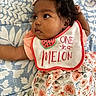 baby, infant, child, bib, bow, curly_hair, floral_pattern, dress, blanket, lying_down, cute, face, expression, indoors, soft_light, patterned_fabric, clothing, head_accessory, skin, person