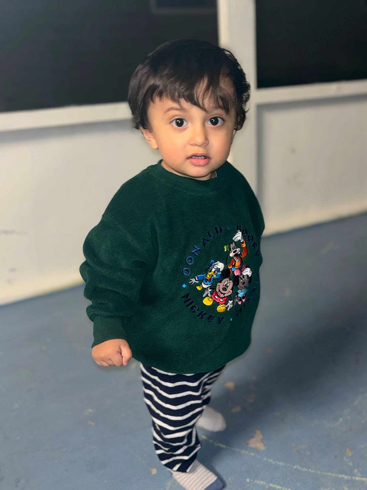 Julius is registered to the contest to win money with this photo: child, toddler, standing, sweatshirt, disney, mickey_mouse, striped_pants, indoor, floor, curious, young_child, casual_clothing, portrait, cute, person, footwear, expression, long_sleeves, hair, background