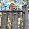 child, toddler, smiling, playground, outdoor, forest, trees, sunny, portrait, face, blonde_hair, pink_clothing, jacket, play_structure, wooden_bars, climbing, happy, park, daylight, photography