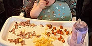 Asher is registered to the contest to win money with this photo: toddler, child, high_chair, bib, food, macaroni, carrots, meat, feeding, indoor, flooring, plastic_bottle, baby_bottle, eating, person, face, hair, feet, wood_floor, dining