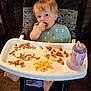 toddler, child, high_chair, bib, food, macaroni, carrots, meat, feeding, indoor, flooring, plastic_bottle, baby_bottle, eating, person, face, hair, feet, wood_floor, dining