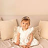 toddler, child, girl, white_dress, headband, barefoot, bed, pillow, beige, cozy, portrait, indoor, cute, smiling, young_child, sitting, soft_lighting, photography, neutral_colors, watermark