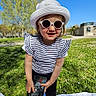 child, girl, sunglasses, hat, smartphone, outdoor, grass, blue_sky, sunny, striped_shirt, blanket, park, casual_clothing, smiling, daylight, person, nature, young_child, fashion, playful