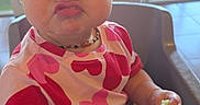 Abriella is registered to the contest to win money with this photo: baby, blue_eyes, child, chubby_cheeks, cucumber, cute, daylight, expression, furniture, heart_pattern, high_chair, indoor, necklace, person, pink_shirt, playful, pucker_face, table, toddler, window