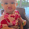 baby, blue_eyes, child, chubby_cheeks, cucumber, cute, daylight, expression, furniture, heart_pattern, high_chair, indoor, necklace, person, pink_shirt, playful, pucker_face, table, toddler, window