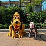 dog, pluto, plush, cartoon_character, outdoor, sunny, harness, greenery, sidewalk, smiling_dog, flower, bush, tree, blue_sky, shadow, concrete, fence, happy, pet, animal