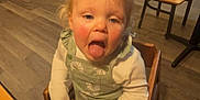 Abigail is registered to the contest to win money with this photo: toddler, child, high_chair, restaurant, wooden_chair, wooden_table, flooring, blonde_hair, tongue_out, long_sleeves, overalls, dining_area, empty_chairs, ceiling_lights, indoor, playful, rosy_cheeks, person, casual_clothing, seat