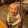 toddler, child, high_chair, restaurant, wooden_chair, wooden_table, flooring, blonde_hair, tongue_out, long_sleeves, overalls, dining_area, empty_chairs, ceiling_lights, indoor, playful, rosy_cheeks, person, casual_clothing, seat