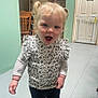 toddler, child, indoor, blonde_hair, pigtails, smiling, happy, standing, long_sleeve_shirt, floral_pattern, black_pants, wooden_chair, kitchen, floor, door, baby_gate, wall, cabinet, light_switch, expression