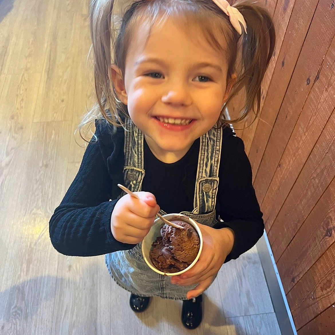 Lilly-Sue joined the competition — help win amazing prizes! black_shirt, bow, child, cup, denim_overalls, dessert, food, girl, happy, holding, ice_cream, indoor, person, pigtails, shoes, smile, spoon, toddler, wooden_floor, wooden_wall