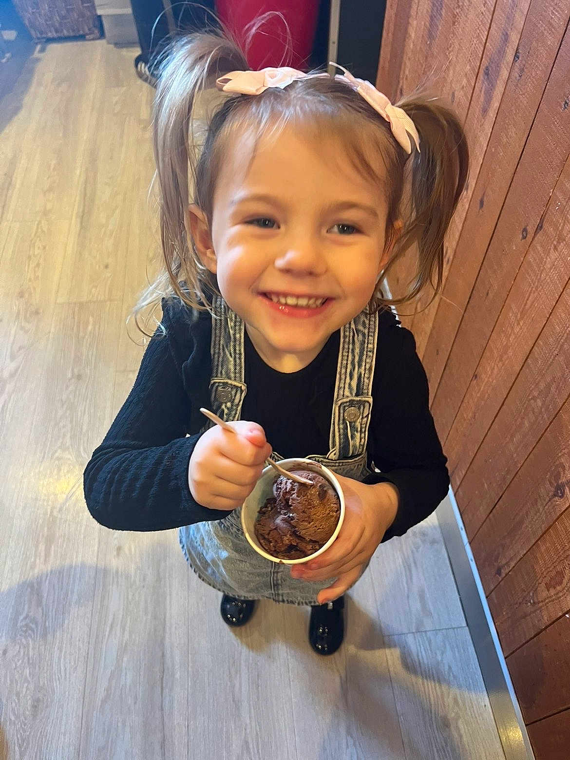 Lilly-Sue joined the competition — help win amazing prizes! child, girl, smile, ice_cream, cup, pigtails, bow, black_shirt, denim_overalls, wooden_floor, wooden_wall, indoor, happy, person, food, dessert, toddler, holding, spoon, shoes