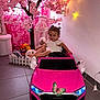 child, toddler, toy_car, pink, indoor, flower_tree, stuffed_animals, fairy_lights, smiling, pigtails, floor_tiles, white_dress, butterfly_sticker, decor, cozy, playtime, steering_wheel, fence, cage, happy